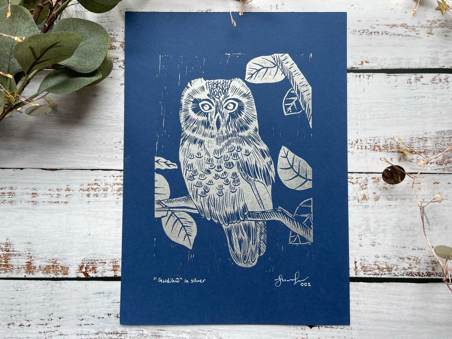 A lino print of an owl sitting in a tree in silver on blue paper